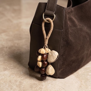 May include: A dark brown suede handbag featuring a decorative bag charm attached to a metal D-ring. The charm consists of a braided beige cord loop adorned with a cluster of smooth dark brown beads and textured light beige seed pods.
