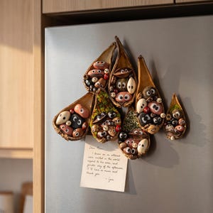 Peut inclure: Un ensemble d'aimants décoratifs composé de six gousses de graines séchées en forme de goutte, remplies de petites pierres peintes avec des yeux. Une note rectangulaire avec du texte manuscrit est fixée en bas. Le texte indique : Dear Poem, I know an m attenus were invited in the world repped to his area, and gender day of the afternoon it'll have so music and thank you. - Grie.