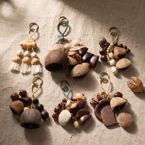 May include: Six decorative hanging ornaments featuring natural elements like dark brown seed pods, smooth wooden beads, and textured nut shells. Each piece is attached to a metal ring with cord, including one with a small beige ceramic cloud.