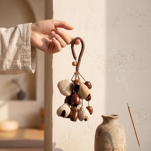May include: A hanging ornament featuring a cluster of dark brown polished seed pods and light beige textured dried fruit shells suspended from a braided brown cord loop with a small wooden bead accent at the top of the central grouping.