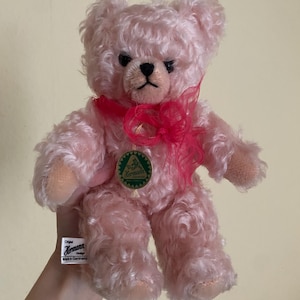 Peut inclure: Un ours en peluche rose clair à poils bouclés, avec des yeux sombres et un nez noir brodé. Il porte un ruban rouge vif transparent autour du cou, une étiquette circulaire verte avec le texte Hermann et une étiquette blanche Original Hermann design Made in Germany.