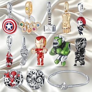 Op de afbeelding: Een verzameling metalen sieradenbedels met superheldenfiguren en symbolen, waaronder een schild, een handschoen, een hamer en een armband met een Avengers-logo. De bedels zijn uitgevoerd in diverse kleuren zoals rood, groen, goud en zilver.