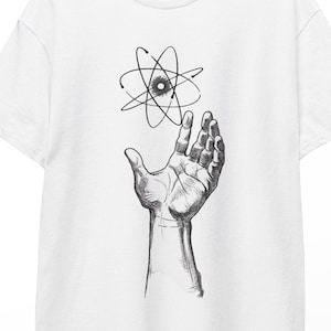 Könnte beinhalten: Ein weißes Kurzarm-T-Shirt mit einer schwarz-weißen Grafikillustration in der Mitte der Brust. Das Design zeigt eine detaillierte menschliche Hand, die nach oben zu einem schwebenden Atomsymbol mit kreisenden Elektronen und einem Kern greift.