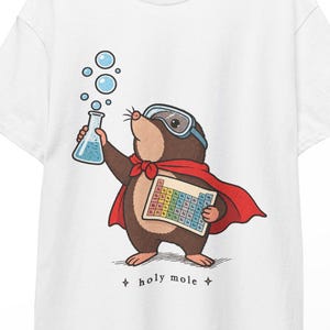 Op de afbeelding: Een wit t-shirt met een afbeelding van een bruine mol die een blauwe veiligheidsbril en een rode cape draagt. De mol houdt een met blauwe vloeistof gevulde beker met bubbels en een periodiek systeem vast. Onder de mol staat de tekst holy mole.