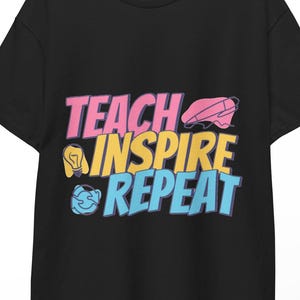 以下が含まれることがあります： ピンク、イエロー、ブルーの文字で「TEACH INSPIRE REPEAT」とプリントされた黒の半袖Tシャツ。デザインにはピンクの鉛筆、黄色の電球、青い地球儀のアイコンが含まれています。Tシャツは白い背景の前で木製のハンガーに掛けられています。