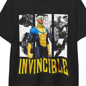 Pode incluir: Uma t-shirt preta de manga curta com uma estampa gráfica da personagem Invincible num fato azul e amarelo. O design inclui três painéis verticais com ilustrações estilo banda desenhada e a palavra INVINCIBLE em letras amarelas abaixo.