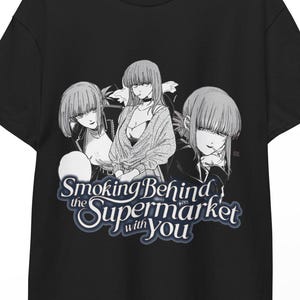 Op de afbeelding: Een zwart t-shirt met korte mouwen voorzien van een grijstinten manga-stijl afbeelding van drie personages, met de tekst "Smoking Behind the Supermarket with You" in een gestileerd wit lettertype met een donkere rand op de borst.