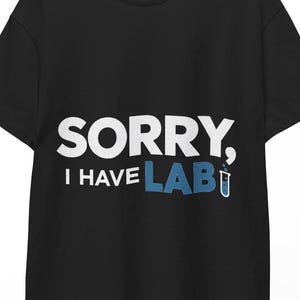 Peut inclure: Un t-shirt noir à manches courtes avec le texte blanc SORRY, I HAVE et le texte bleu LAB, où la lettre B est remplacée par un petit graphique de tube à essai bleu. Le t-shirt est présenté sur un cintre en bois sur un fond blanc.