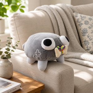 May include: A round, gray plush toy with large white and black eyes, four short legs, and a small embroidered flower on its side. The plush toy holds a cream-colored bone-shaped object tied with a small, vibrant rainbow-colored ribbon.