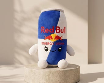May include: A plush toy shaped like a Red Bull energy drink can. The soft fabric features the blue and white colour scheme, the Red Bull logo, and the text "Red Bull ENERGY DRINK." It has small white arms and legs and a stitched smiling face.