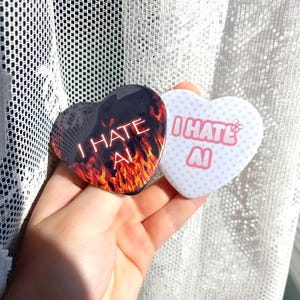 May include: Two heart-shaped pin-back buttons. One button features a black background with orange and red flames and the white text I HATE AI. The second button is white with light pink polka dots and the pink text I HATE AI with a small butterfly.