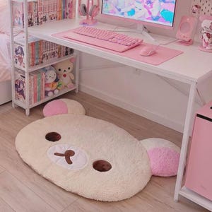 May include: A plush, oval-shaped floor rug designed to resemble a bear face. The rug features a cream-coloured fuzzy texture, dark brown embroidered eyes and nose, a white muzzle area, and two rounded ears with light pink accents on the top.