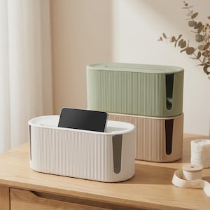 Op de afbeelding: Twee ovale kabelmanagementboxen met verticaal geribbelde texturen. Een witte doos bevat een zwarte smartphone in de bovenste gleuf, terwijl een saliegroene doos op een beige basiseenheid is gestapeld, beide voorzien van uitsparingen aan de zijkanten.