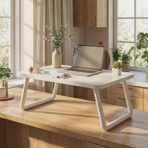 Puede incluir: Un escritorio portátil blanco para computadora con bordes redondeados y patas en forma de U. La superficie sostiene una computadora plateada, un libro pequeño abierto, un jarrón beige con ramas de eucalipto y una suculenta con auriculares.