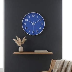 May include: A round wall clock with a vibrant blue face, white Arabic numerals, and thin white hands. The clock features a simple white circular frame. It is mounted on a dark grey wall above a wooden shelf holding a vase with dried grass.