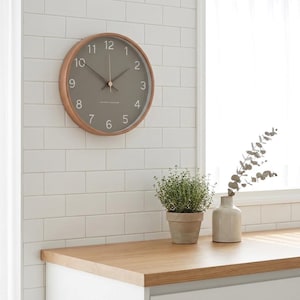 Op de afbeelding: Een ronde wandklok met een lichthouten frame en een matgrijze wijzerplaat. De klok is voorzien van witte Arabische cijfers van één tot twaalf, dunne witte uur- en minutenwijzers en een dunne witte secondewijzer. In het midden staat kleine witte tekst.
