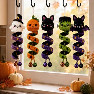 May include: Five hanging crochet ornaments featuring Halloween characters: a white ghost with a hat, an orange pumpkin, a black cat, a green monster, and a black bat. Each figure has a long, spiraling tail decorated with stars and two small metal bells.