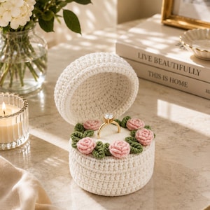 May include: A round, cream-colored crocheted ring box with an open lid. The interior features a soft white cushion decorated with small, pink crocheted roses and green leaves. A gold ring with a clear stone is centered in the cushion.