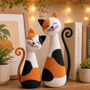 Puede incluir: Dos figuras de gato tejidas a ganchillo a mano con un patrón de calicó en hilo blanco, naranja y negro. Cada gato tiene un cuerpo esbelto, una cola enroscada y una cara bordada con ojos cerrados, nariz rosa y bigotes negros.
