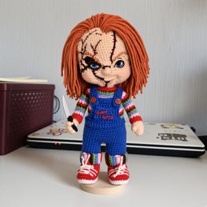 May include: A crocheted doll with orange yarn hair, featuring a scarred face, blue eyes, and a striped long-sleeve shirt under blue overalls with the text Good Guys embroidered in red. The doll holds a small white knife and wears red sneakers.