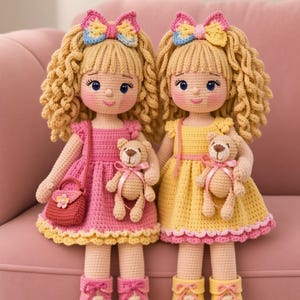 May include: Two handmade crochet dolls with curly blonde yarn hair, wearing colorful dresses and matching bows. One doll wears a pink dress with a small purse, the other a yellow dress. Both dolls hold a small beige crochet teddy bear with a pink ribbon.