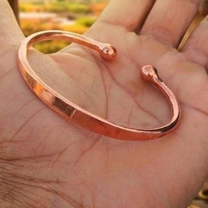 May include: A polished copper cuff bracelet with a smooth, flat band and rounded ball terminals at each end. The bracelet features an open design, allowing for adjustment, and displays a warm, metallic reddish-brown colour throughout the surface.