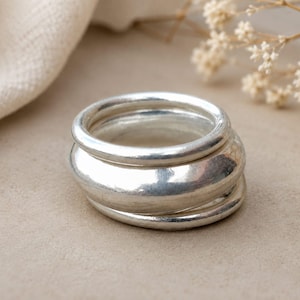 Puede incluir: Un conjunto de tres anillos de plata con un acabado liso y pulido. Los anillos varían en grosor, con una banda más ancha y redondeada situada entre dos bandas circulares más finas, creando una apariencia superpuesta con una superficie metálica.