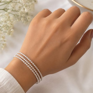 Può includere: Un set di tre braccialetti sottili color argento in stile corda indossati al polso. Ogni braccialetto presenta una texture metallica ritorta continua che crea un motivo sottile e riflettente. I braccialetti sono indossati insieme come set.