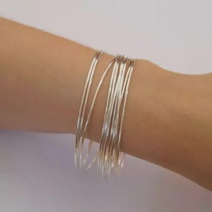 Puede incluir: Un conjunto de múltiples brazaletes finos de metal en tono plateado pulido que se usan juntos. Las pulseras son circulares, lisas y reflectantes, creando una apariencia apilada en la muñeca. Cada brazalete tiene un grosor uniforme.