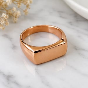 Puede incluir: Un anillo de sello en oro rosa con un acabado liso y pulido y una cara plana rectangular. La banda es sólida y de ancho uniforme, con un tono metálico cálido que refleja la luz a través de su superficie curva y su parte superior.