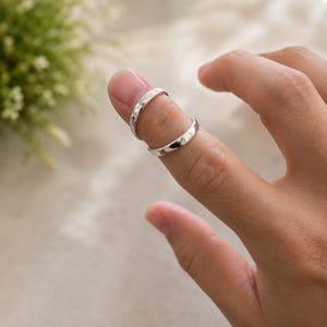Könnte beinhalten: Zwei polierte silberfarbene Metallringe mit glattem, abgerundetem Banddesign. Die Ringe sind an einem Finger positioniert, wobei einer in der Nähe des Knöchels und der andere weiter unten in Richtung Fingerbasis sitzt und eine reflektierende Oberfläche zeigt.
