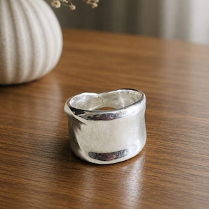 Puede incluir: Un anillo ancho de plata con una forma orgánica irregular y una superficie metálica lisa y reflectante. La banda presenta un borde grueso y ondulado que crea una silueta no uniforme, mostrando un acabado pulido que refleja la luz.