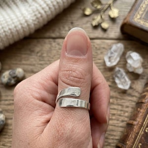 Può includere: Un anello color argento con un design aperto in stile bypass caratterizzato da una finitura liscia e lucida. La fascia è composta da due estremità piatte e arrotondate che si sovrappongono attorno al dito, creando un aspetto semplice e pulito.