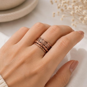 May include: A wide copper-toned ring featuring a central twisted rope-style band flanked by two outer bands with an embossed floral pattern. The metal has a warm, reddish-brown finish with darkened recessed areas that highlight the intricate details.