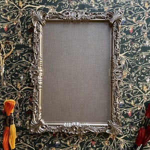 May include: A rectangular display board featuring a metallic silver-toned frame with intricate, ornate scrollwork and floral relief detailing. The centre of the frame is filled with a fine, uniform grid mesh screen, providing a structured surface.
