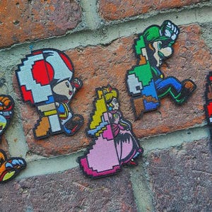 Puede incluir: Un conjunto de cinco parches bordados con personajes de videojuegos pixelados, incluyendo a Bowser, Toad, la Princesa Peach, Luigi y Mario. Cada parche muestra colores vibrantes y contornos en bloque típicos de los gráficos de 8 bits.