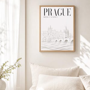 May include: A framed wall art print featuring the word PRAGUE in bold black capital letters above geographic coordinates and a white embossed-style illustration of the Prague skyline, including the Charles Bridge and castle architecture.