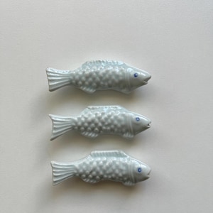 May include: Three ceramic chopstick rests shaped like fish, each featuring a glossy white finish with a textured scale pattern and a small blue dot for the eye. The fish are arranged vertically in a row, showcasing a uniform design and shape.