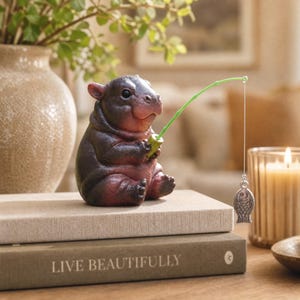 May include: A small, dark brown and reddish-toned hippo figurine sitting upright. The hippo holds a thin, bright green fishing rod with a silver-colored metal fish charm dangling from a white string, creating a playful and detailed decorative ornament.