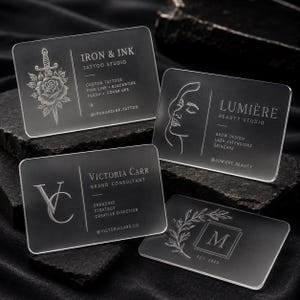 May include: Four rectangular frosted acrylic business cards with rounded corners. Each card features white printed text and graphics, including logos for Iron & Ink, Lumière Beauty Studio, Victoria Carr Brand Consultant, and a monogram M with a leaf design.