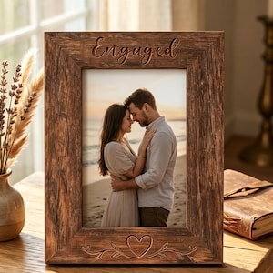 Op de afbeelding: Een rechthoekige houten fotolijst met een donkerbruine, verweerde afwerking. De bovenrand is gegraveerd met het woord "Engaged" in cursief schrift en de onderrand bevat een gegraveerd hart omringd door delicate rankmotieven.