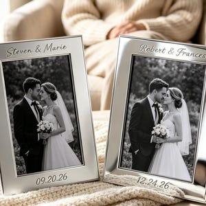 May include: Two rectangular silver-toned metal picture frames featuring engraved text. The left frame is engraved with "Steven & Marie" and "09.20.26". The right frame is engraved with "Robert & Frances" and "12.24.26". Each frame holds a black and white wedding photo.