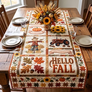 May include: A rectangular quilted table runner featuring autumn motifs including a scarecrow, a red tractor, pumpkins, sunflowers, acorns, and fall leaves. The text HELLO FALL is embroidered in brown. The runner is centered on a wooden dining table.