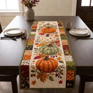 Può includere: Un runner da tavola rettangolare con un pannello centrale raffigurante quattro zucche impilate nei colori arancione, bianco e verde, circondate da foglie autunnali e bacche. Il bordo presenta un design patchwork con vari motivi autunnali.