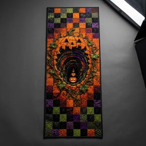 May include: A rectangular quilted table runner featuring a checkered pattern in orange, purple, black, and green. The center displays a large carved pumpkin design with a deep tunnel effect, surrounded by green vines and two smaller jack-o'-lanterns.