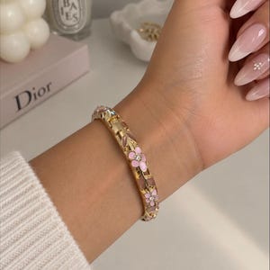 Peut inclure: Un bracelet jonc articulé doré orné d'un motif floral répétitif avec des pétales en émail rose clair et de petits cristaux transparents sertis au centre de chaque fleur, fixé par un mécanisme de fermoir métallique sur le côté du poignet.