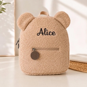 Peut inclure: Un sac à dos en forme d'ours en peluche beige, fabriqué dans un tissu sherpa doux et duveteux. Le sac présente deux oreilles arrondies sur le dessus, une poche frontale zippée avec une étiquette ronde marron et le prénom Alice brodé en noir.