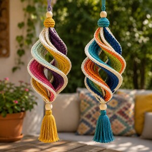 May include: Two hanging spiral ornaments crafted from crocheted yarn in vibrant multi-colored segments. Each piece features a central twisted design with cream-colored edges, topped with a wooden bead and finished with a long, solid-colored tassel.