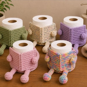 Peut inclure: Cinq rouleaux de papier toilette dans des housses au crochet. Chaque housse présente un design en forme de livre avec de petits bras et jambes. Les housses sont vert olive, beige moucheté, violet, rose et en fil pastel multicolore.