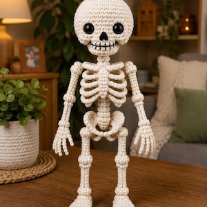 May include: A cream-coloured crocheted skeleton figurine featuring a round skull with large black plastic eyes, a stitched black nose, and a stitched black mouth. The figure has a visible rib cage, articulated joints, and detailed hand and foot structures.